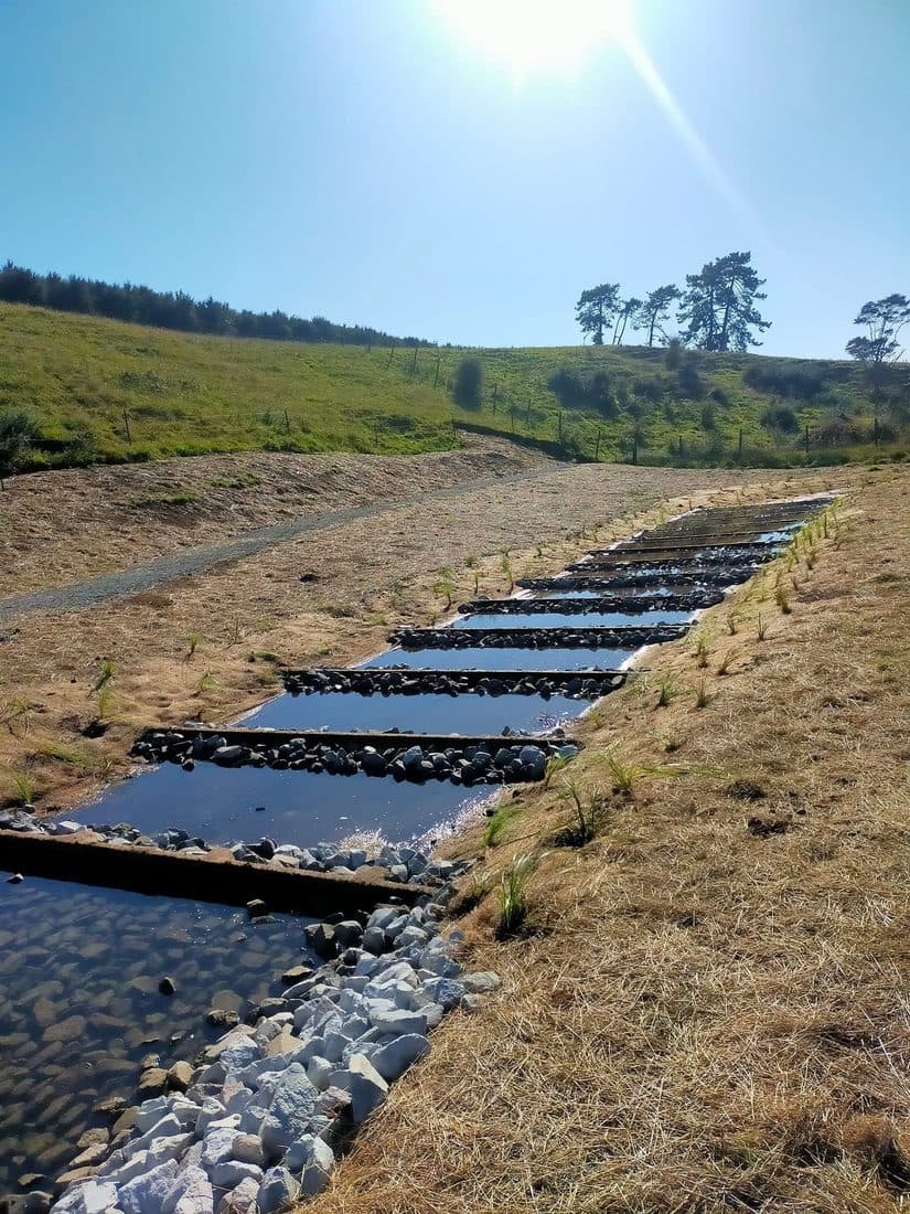 Rock-lined drainage channel with concrete slab crossings running down a grassed hillside